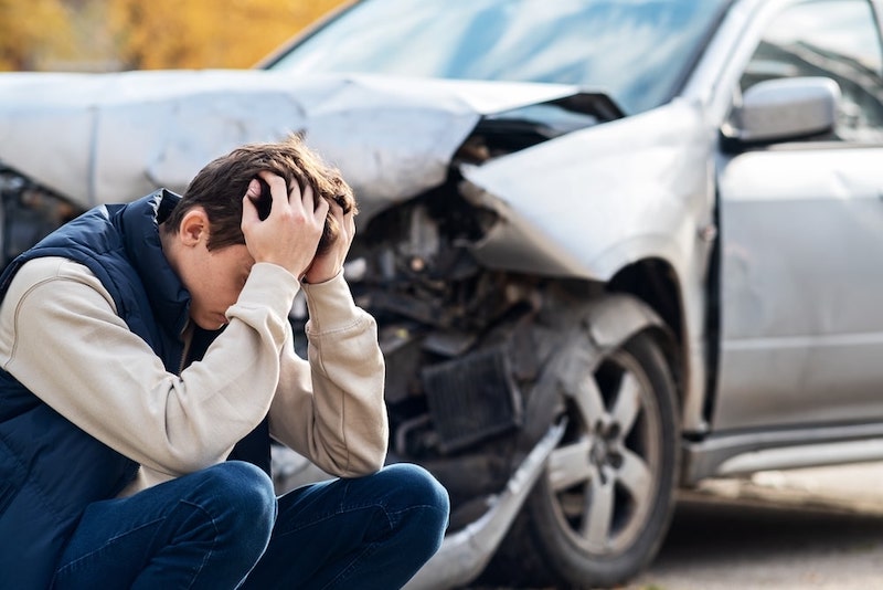 A Boy Holding His Head Next To A Damaged Car